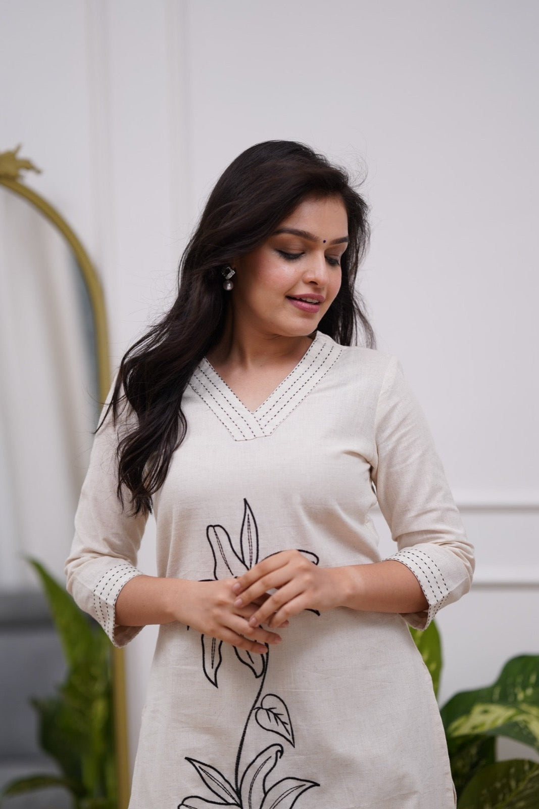 Woman wearing a white dress with black floral embroidery in a minimal indoor setting.